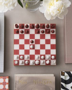 Overhead view of a red-and-white chessboard with red pieces on the top and white pieces on the bottom, beside books and a vase of white flowers.