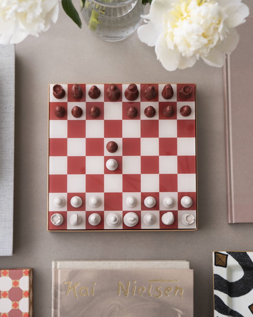 Overhead view of a red-and-white chessboard with red pieces on the top and white pieces on the bottom, beside books and a vase of white flowers.