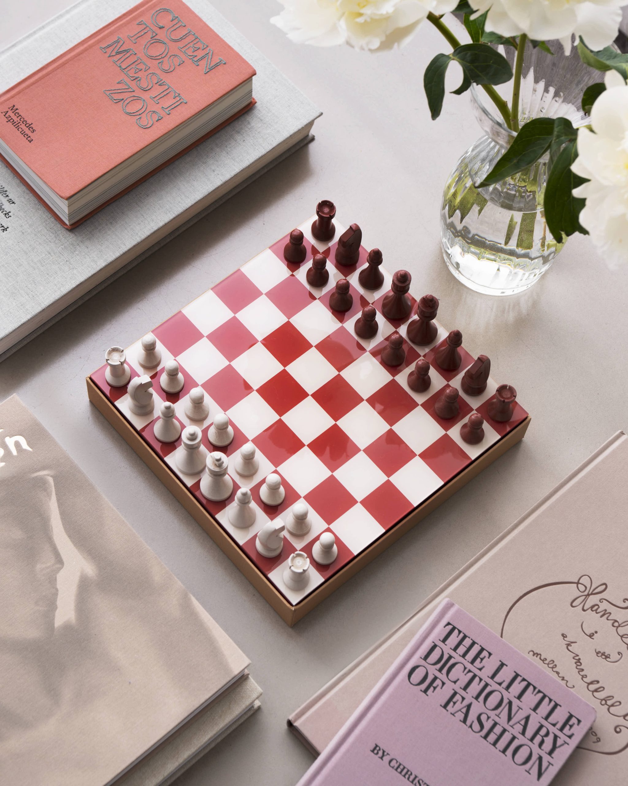 Image 186 - New Trend Flat lay: pink and white checkered chessboard with red/brown chessmen, beside a pink book and a vase of white flowers.