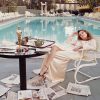 Woman in a cream satin dress lounging by a hotel pool, with newspapers scattered on the tiled deck and a breakfast table nearby.