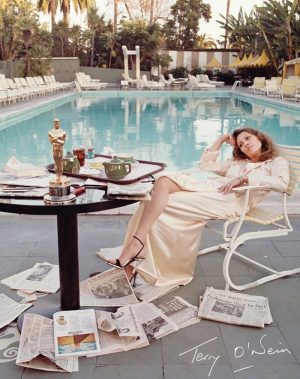 Woman in a cream satin dress lounging by a hotel pool, with newspapers scattered on the tiled deck and a breakfast table nearby.