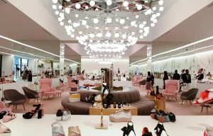 Spacious luxury boutique with pink seating, display tables of shoes, and a central circular ottoman under a chandelier.