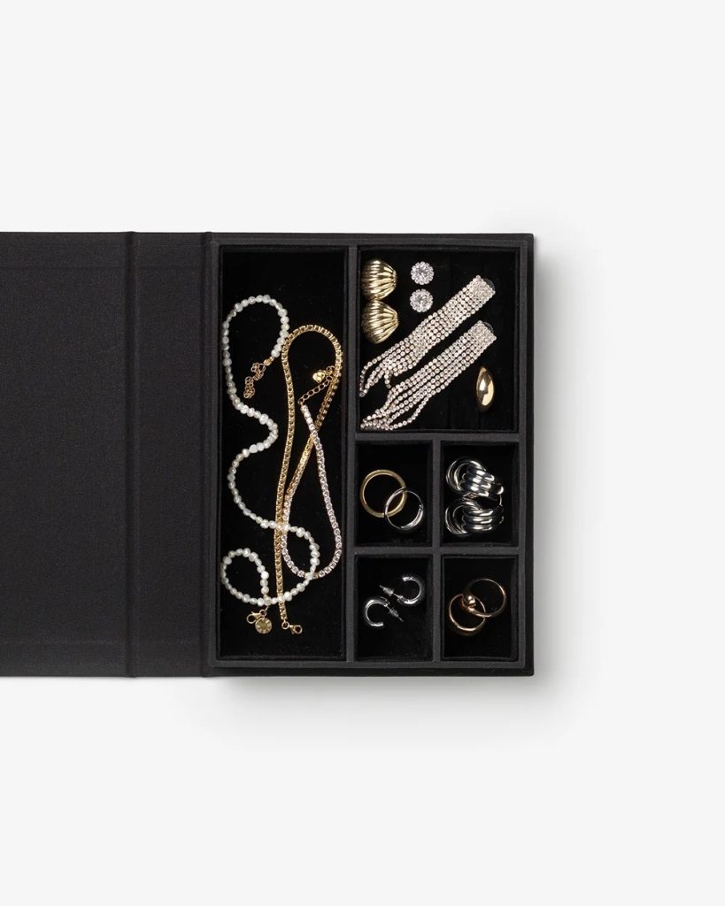 Black jewelry box open to reveal organized compartments with necklaces, rings, bracelets, and earrings in gold, silver, and pearl styles.