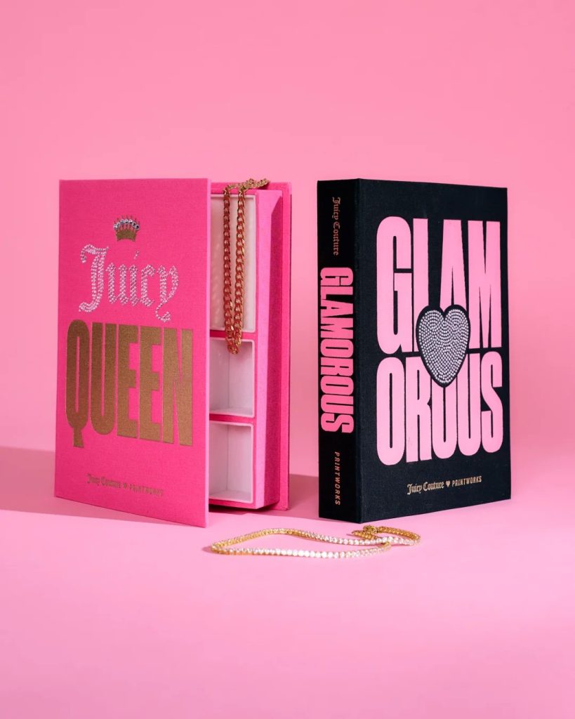 Pink open jewelry box with gold chain necklace displayed; bold 'QUEEN' text on cover beside a black 'GLAMOROUS' book and heart graphic, on pink backdrop.