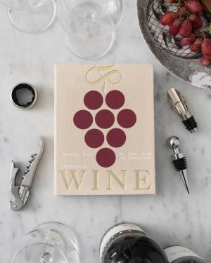 Flat lay of a beige wine book titled 'WINE' with a grape cluster illustration, surrounded by wine accessories on a marble surface.