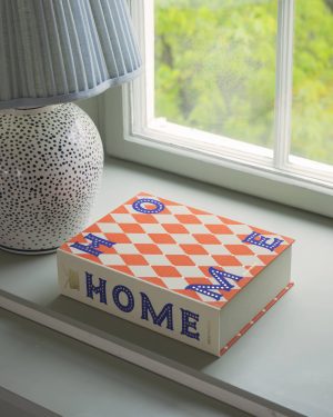 Blue striped lampshade on a windowsill beside a white polka-dotted vase and a red-and-white geometric box with the word 'HOME' in blue letters.
