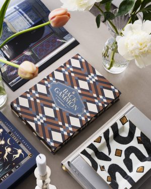 Top-down view of patterned game boxes and a glass vase with white flowers on a neutral tabletop.
