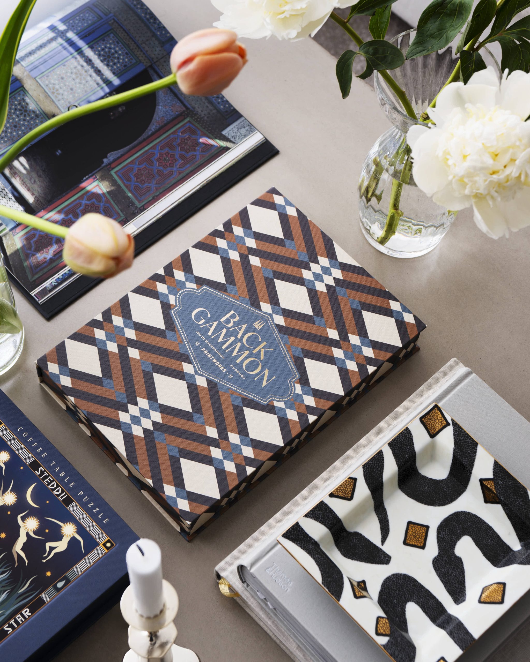 Image 207 - New Trend Top-down view of patterned game boxes and a glass vase with white flowers on a neutral tabletop.