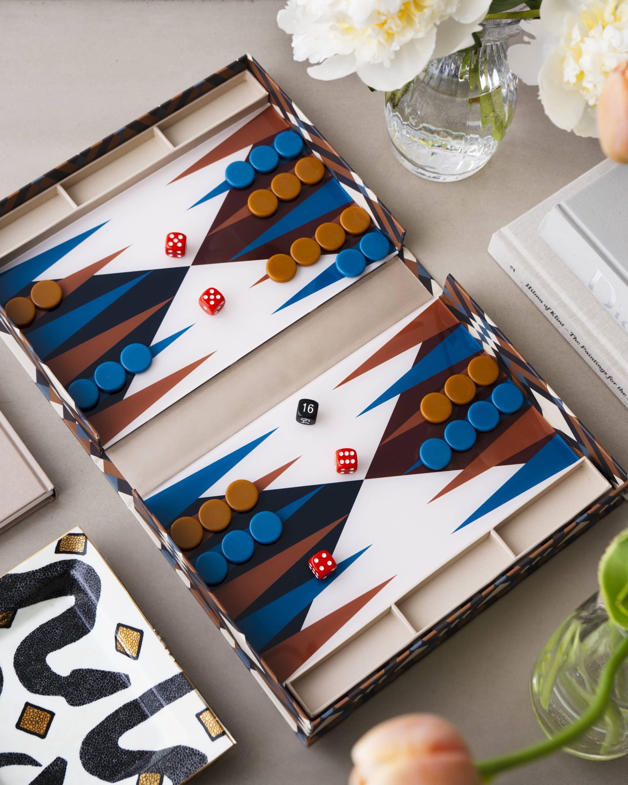 Image 210 - New Trend Overhead view of a backgammon-like board with blue and orange discs and red dice, placed inside a patterned box near a glass vase of white flowers and a closed book or binder.