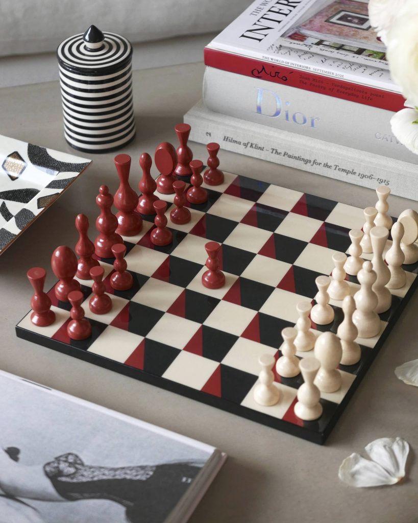 Chessboard with red and white chess pieces arranged for a game, magazines stacked behind, and a black-and-white striped jar on the left.