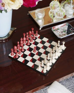 Chessboard with red and white pieces arranged for a game on a dark wooden table, background shows a vase of flowers and a mirrored tray with glassware.