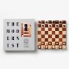 Gray book titled 'The Modernist' next to a wooden chessboard with all pieces in starting positions on a white background.