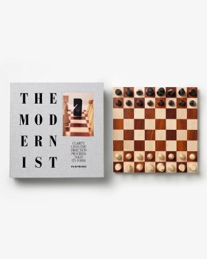 Gray book titled 'The Modernist' next to a wooden chessboard with all pieces in starting positions on a white background.