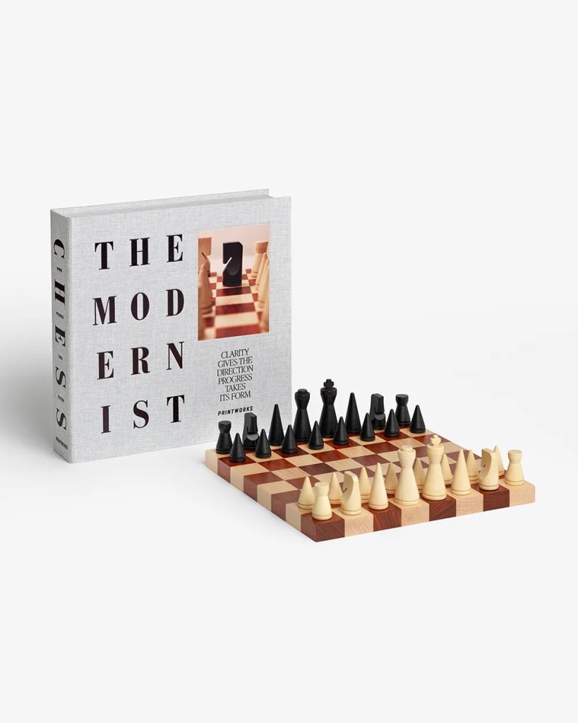Wooden chessboard with black and white pieces set up beside a light-gray hardcover book titled 'The Modernist' on a white background.