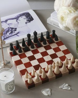 Chessboard with black and white pieces arranged for a game, beside a bouquet of white flowers and a stack of books on a desk.