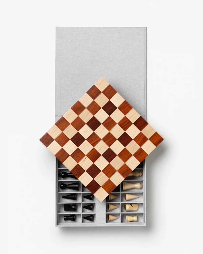 Gray storage box with compartments holding black and white chess pieces; a wooden checkered board rests diagonally across the top.