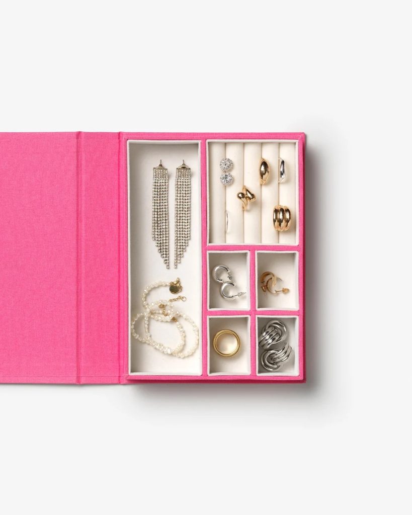 Open pink jewelry box with multiple compartments displaying earrings, rings, and bracelets in silver and gold tones.