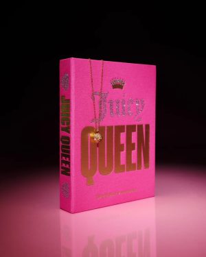 Bright pink hardcover book titled 'QUEEN' with a gold necklace draped over the cover, reflected on a glossy pink surface in a dark studio.