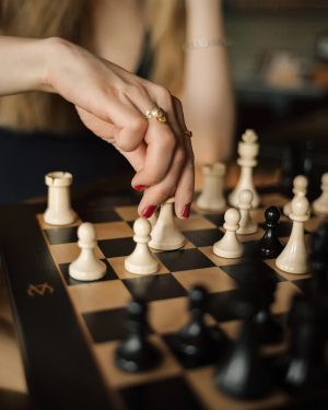 Close-up of a hand with rings moving a white pawn on a chessboard.