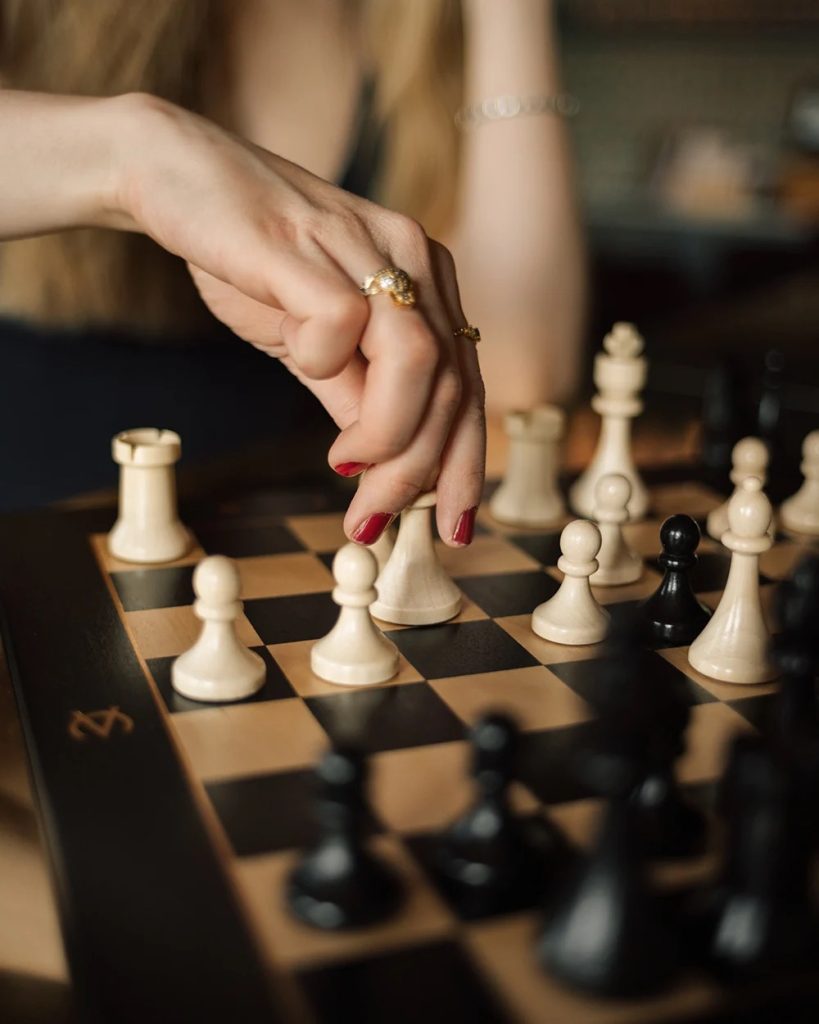 Close-up of a hand with rings moving a white pawn on a chessboard.