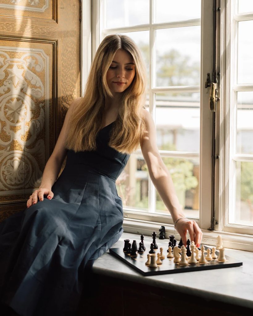 Young woman in a dark gown sits on a windowsill, moving a chess piece on a board by daylight?