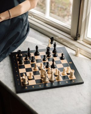 Chessboard on a windowsill with white and black pieces midgame; person in dark clothing sits nearby.