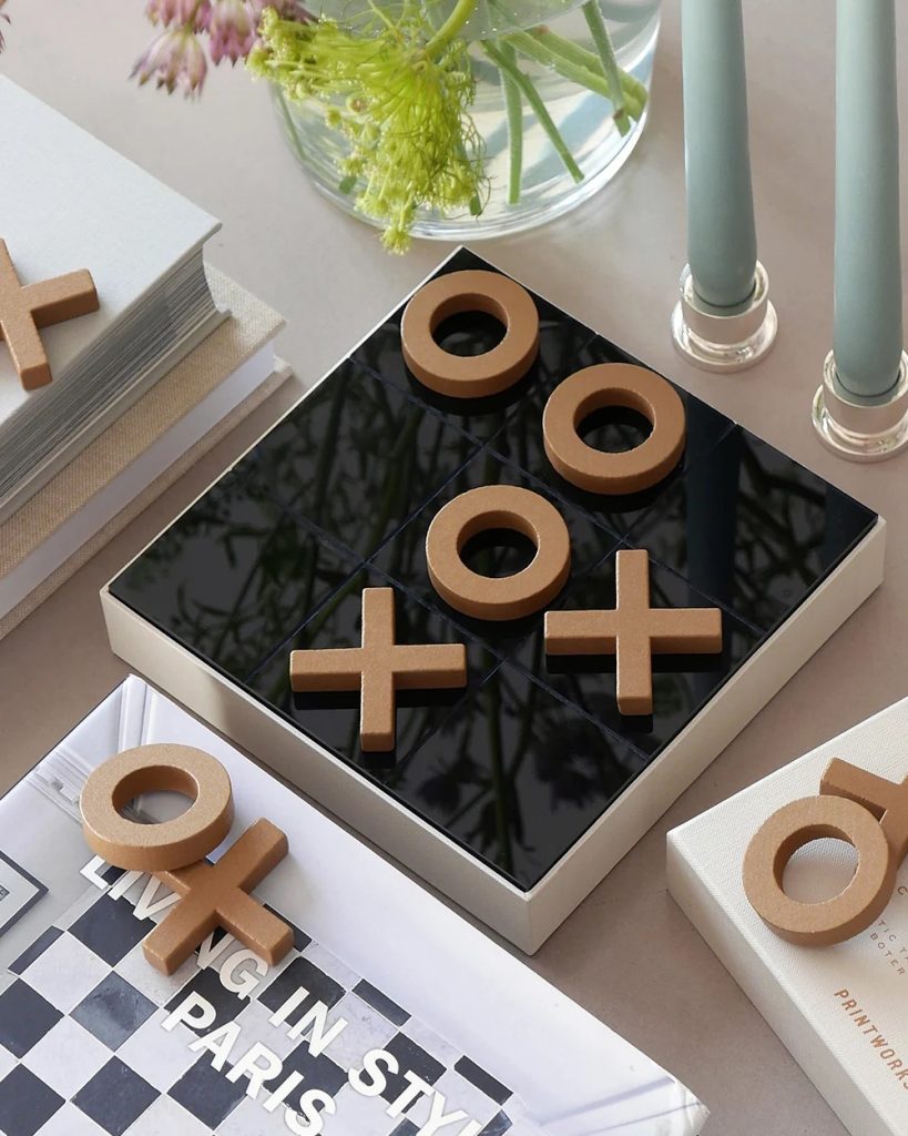 Brown wooden X and O pieces sit on a glossy black tic-tac-toe board, surrounded by a book, flowers, and candles on a neutral desk.