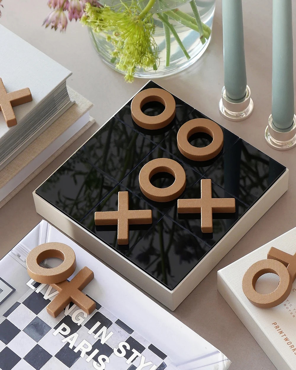 Image 240 - New Trend Brown wooden X and O pieces sit on a glossy black tic-tac-toe board, surrounded by a book, flowers, and candles on a neutral desk.
