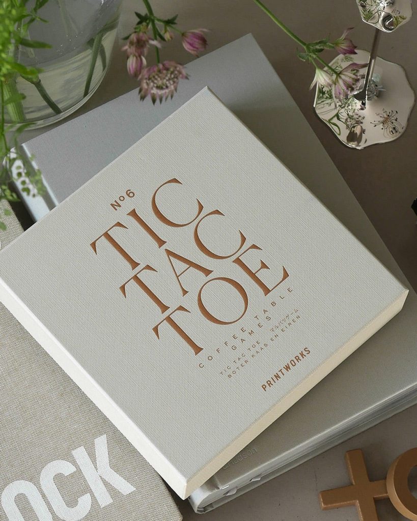 White textured book titled 'No.6 Coffee Table' in copper letters on a table with flowers and a decorative lamp nearby.