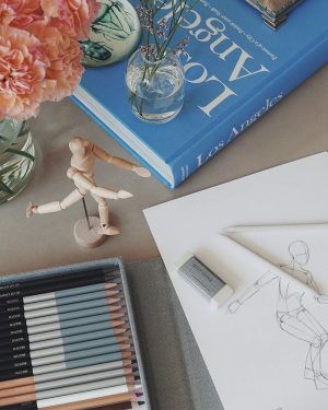 Desklab with pink flowers in a vase, a blue Los Angeles book, a wooden artist mannequin, a set of pencils, eraser, and a fashion sketch on white paper.