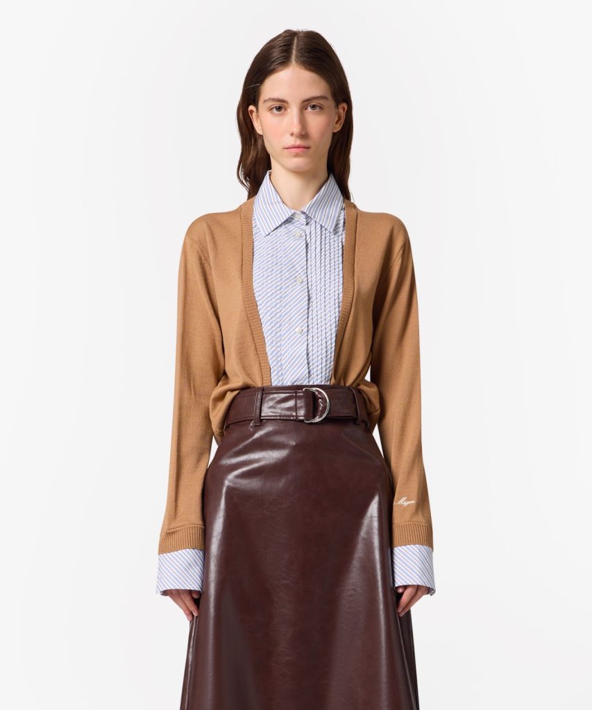 Model wearing a camel cardigan over a blue striped button-down shirt and a brown leather midi skirt with a belt.