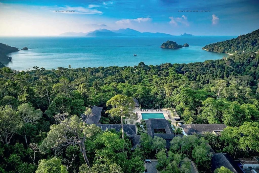 Aerial view of a jungle resort with a central rectangular pool and thatched-roof buildings, overlooking a turquoise sea and distant islands.
