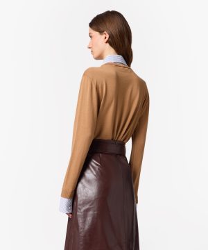 Back view of a model in a camel sweater layered over a striped shirt, tucked into a brown leather midi skirt with a belt.