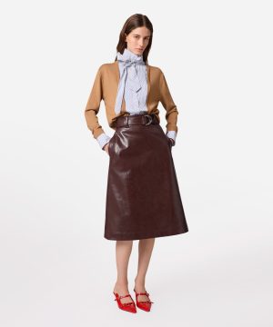Woman in a camel sweater, striped blouse with a large bow, and a brown leather midi skirt with a belt, red shoes.