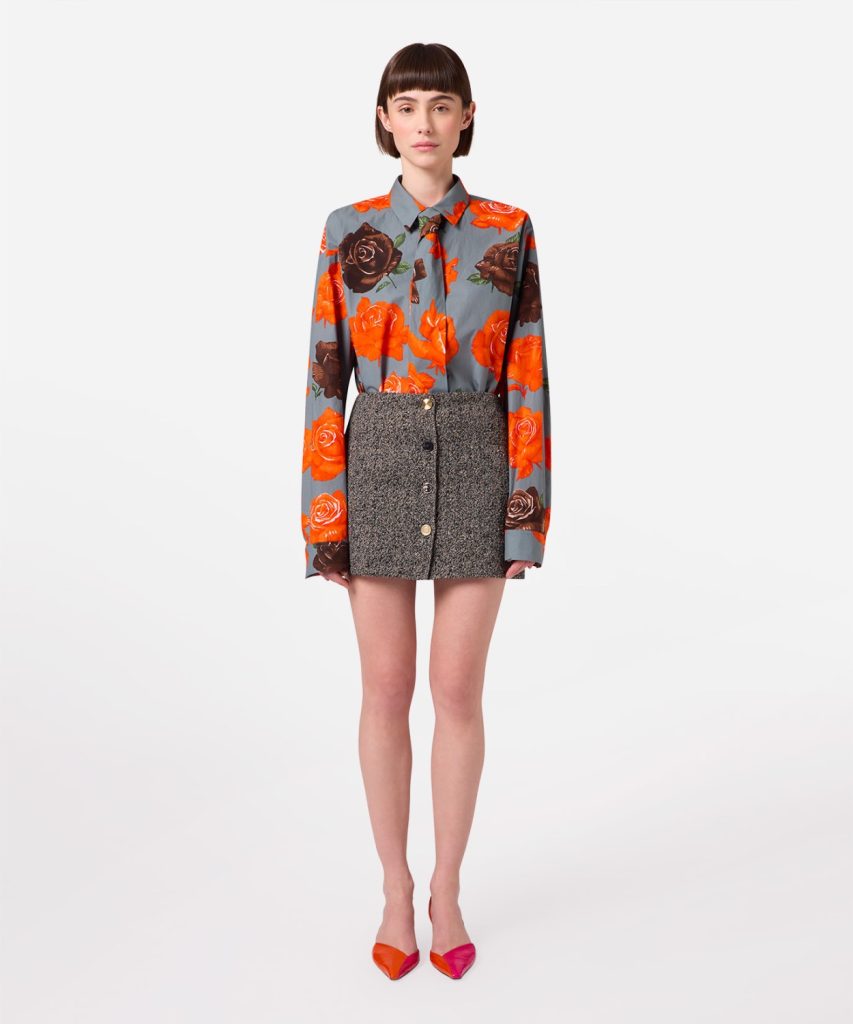 Model wearing a blue-gray floral blouse with orange roses tucked into a textured gray mini skirt with a button front, standing against a white background with pinkish-orange heels nearby.