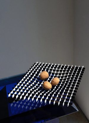 Three walnuts rest on a tilted black-and-white checkered panel in a blue-lit corner of a gallery (abstract sculpture).