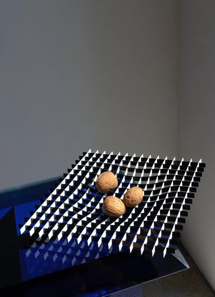 Three walnuts rest on a tilted black-and-white checkered panel in a blue-lit corner of a gallery (abstract sculpture).