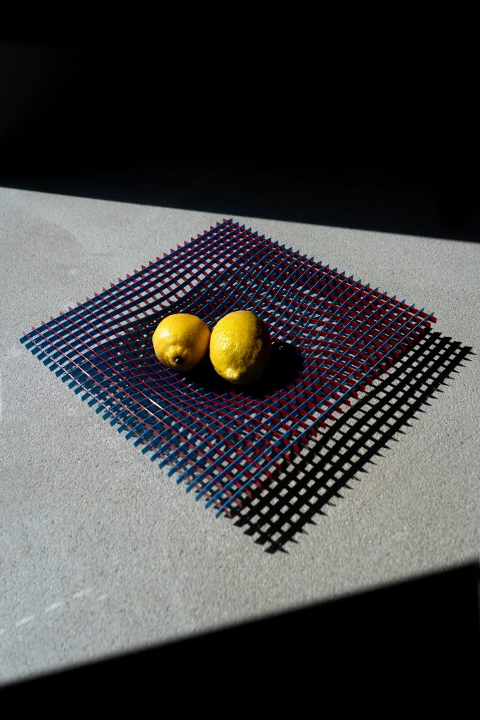 Two lemons sit on a blue-and-red perforated mat cast in bright light on a concrete surface, creating strong shadows.