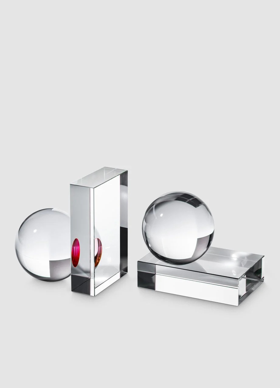 Image 265 - New Trend Three transparent glass objects: a sphere on the left, a tall rectangular prism with a pink accent, and a sphere resting on a rectangular base to the right, all on a light gray background.