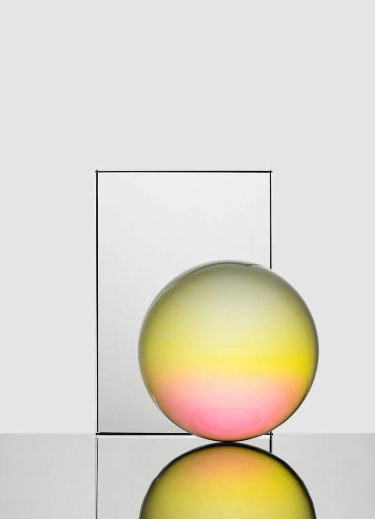 Glossy glass sphere with a yellow-to-pink gradient sits beside a tall rectangular frame, reflected on a glossy surface.