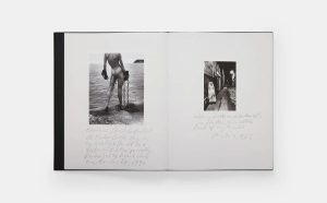 Open book showing two black-and-white photographs with handwritten captions on white pages; left image depicts a nude figure by the water with chains, right image shows a street scene with a figure in a coat.