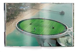 Aerial view of a circular rooftop tennis court perched over blue-green water, with players on the green surface and a beach below.