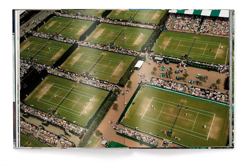 Aerial view of several grass tennis courts inside a stadium, with players competing and large crowds watching from stands.