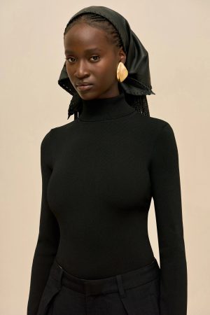 Portrait of a woman in a black turtleneck with a black headscarf and large gold earrings against a beige background
