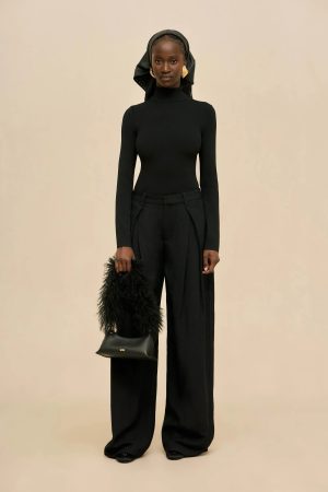 Model wearing a fitted black turtleneck and wide-leg trousers with a black headscarf, standing against a beige backdrop.