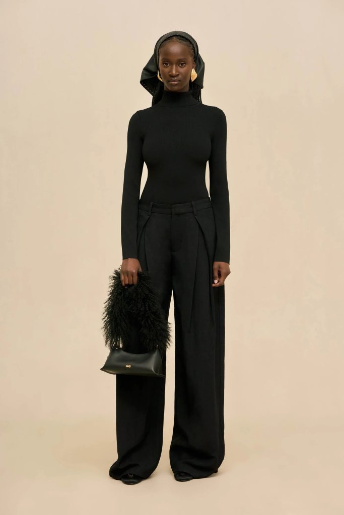 Model wearing a fitted black turtleneck and wide-leg trousers with a black headscarf, standing against a beige backdrop.