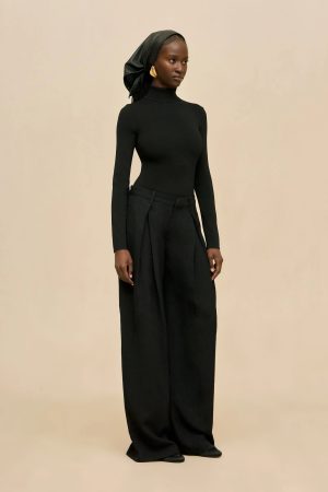 Woman in a black turtleneck and wide-leg black pants, accessorized with a black headscarf and gold earrings, stands against a beige background.