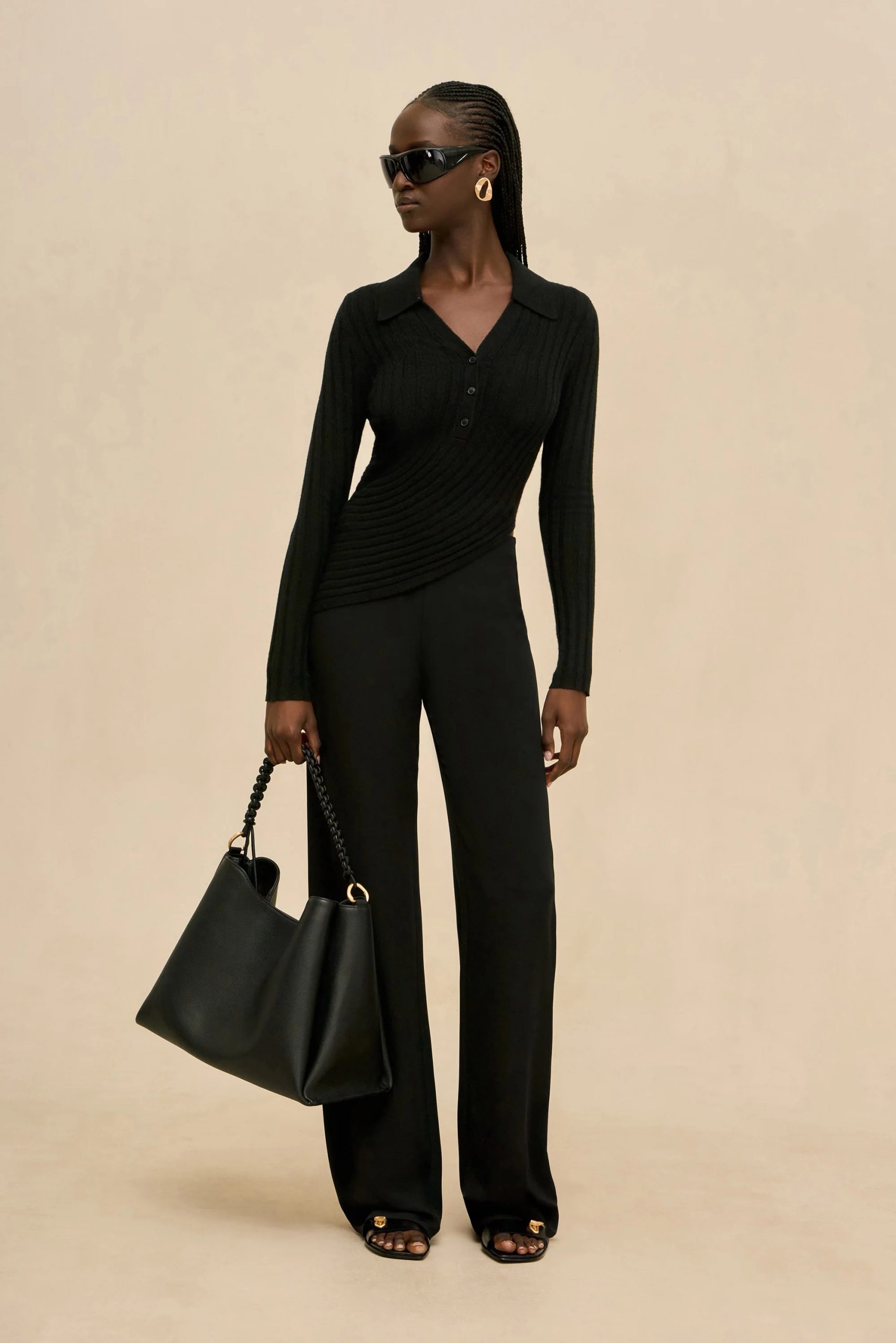 Image 409 - New Trend Woman in an all-black outfit (ribbed long-sleeve top and wide-leg pants) wearing oversized sunglasses and gold earrings, carrying a black handbag with a chain strap against a beige backdrop.