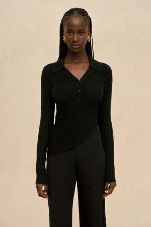 Dark-skinned woman in a black ribbed knit top with a V-neck and buttons, wearing gold hoop earrings, standing against a beige background.