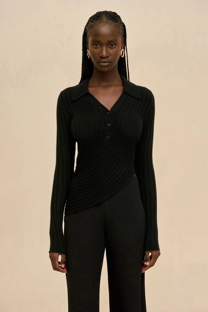 Dark-skinned woman in a black ribbed knit top with a V-neck and buttons, wearing gold hoop earrings, standing against a beige background.